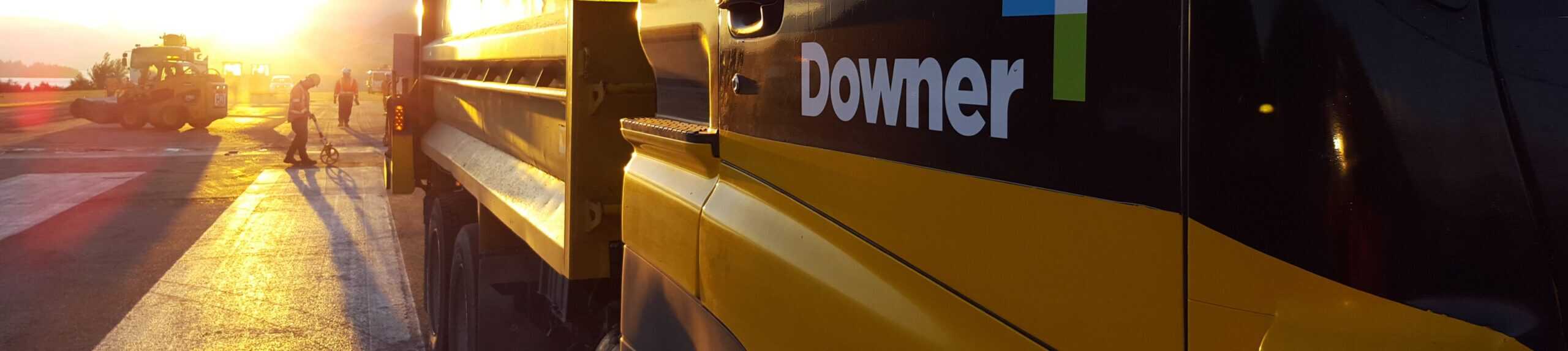 Construction workers in safety gear measure and work on an airport runway at sunrise, with golden light reflecting off the asphalt. A large yellow Downer vehicle is in the foreground, and heavy machinery and workers are visible in the distance.