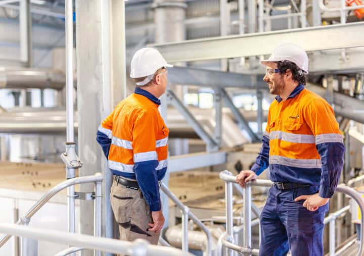 Two workers wearing orange high-visibility uniforms, safety helmets, and protective glasses are standing on a raised platform inside an industrial facility. They are engaged in conversation, surrounded by metal railings, pipes, and machinery, suggesting a manufacturing or processing plant environment.