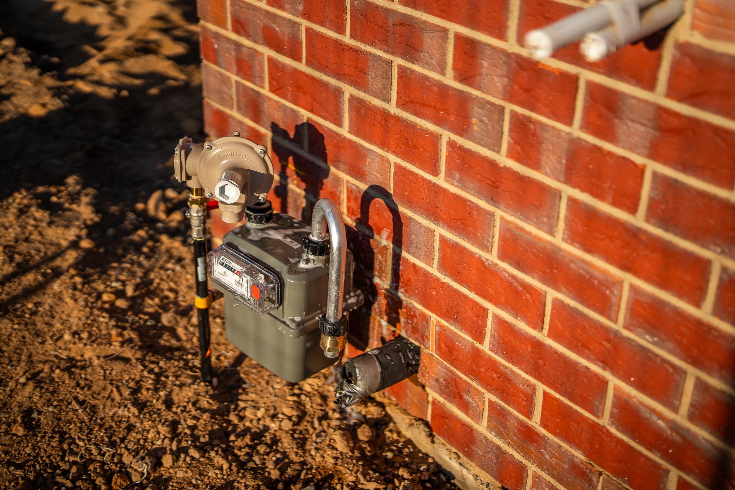 A close-up, outdoor shot shows a residential gas meter and pressure regulator installed on the exterior of a newly constructed building. The wall is made of red brick with white mortar. The gas meter is a dark gray box with a digital readout and is mounted at the bottom of the wall. A lighter gray gas regulator is connected to the meter via pipes and fittings. The ground around the installation is dry, reddish-brown soil and gravel, and the late afternoon sun casts long shadows.