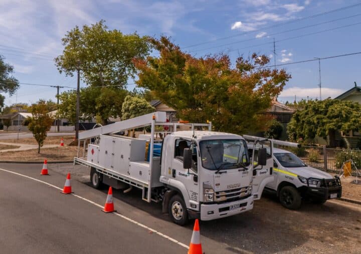 A sunny outdoor image showing two Downer utility vehicles parked on a suburban street. In the foreground is a white Isuzu utility truck with a flatbed body and toolboxes, along with an adjacent white and yellow utility pickup truck. Both vehicles are parked on the side of an asphalt road with a line of orange safety cones extending from the foreground. Behind the trucks are established suburban houses and trees showing early autumn colors. The sky is blue with some white clouds.