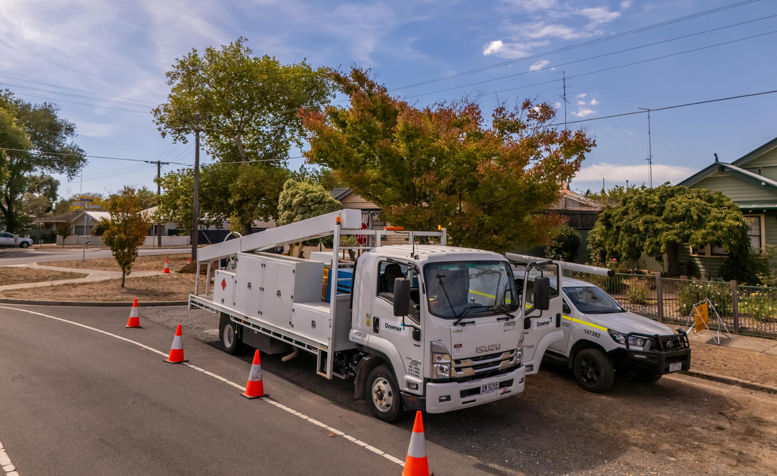 A sunny outdoor image showing two Downer utility vehicles parked on a suburban street. In the foreground is a white Isuzu utility truck with a flatbed body and toolboxes, along with an adjacent white and yellow utility pickup truck. Both vehicles are parked on the side of an asphalt road with a line of orange safety cones extending from the foreground. Behind the trucks are established suburban houses and trees showing early autumn colors. The sky is blue with some white clouds.