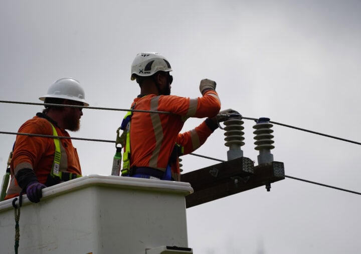 An outdoor, slightly low-angle shot shows two utility workers in a bucket truck, working on overhead power lines against a cloudy sky. Both wear safety harnesses, hard hats (one white, one dark), and high-visibility orange shirts. The worker on the right is actively handling the wires near porcelain insulators mounted on a wooden crossarm. The worker on the left is looking toward the center of the frame.