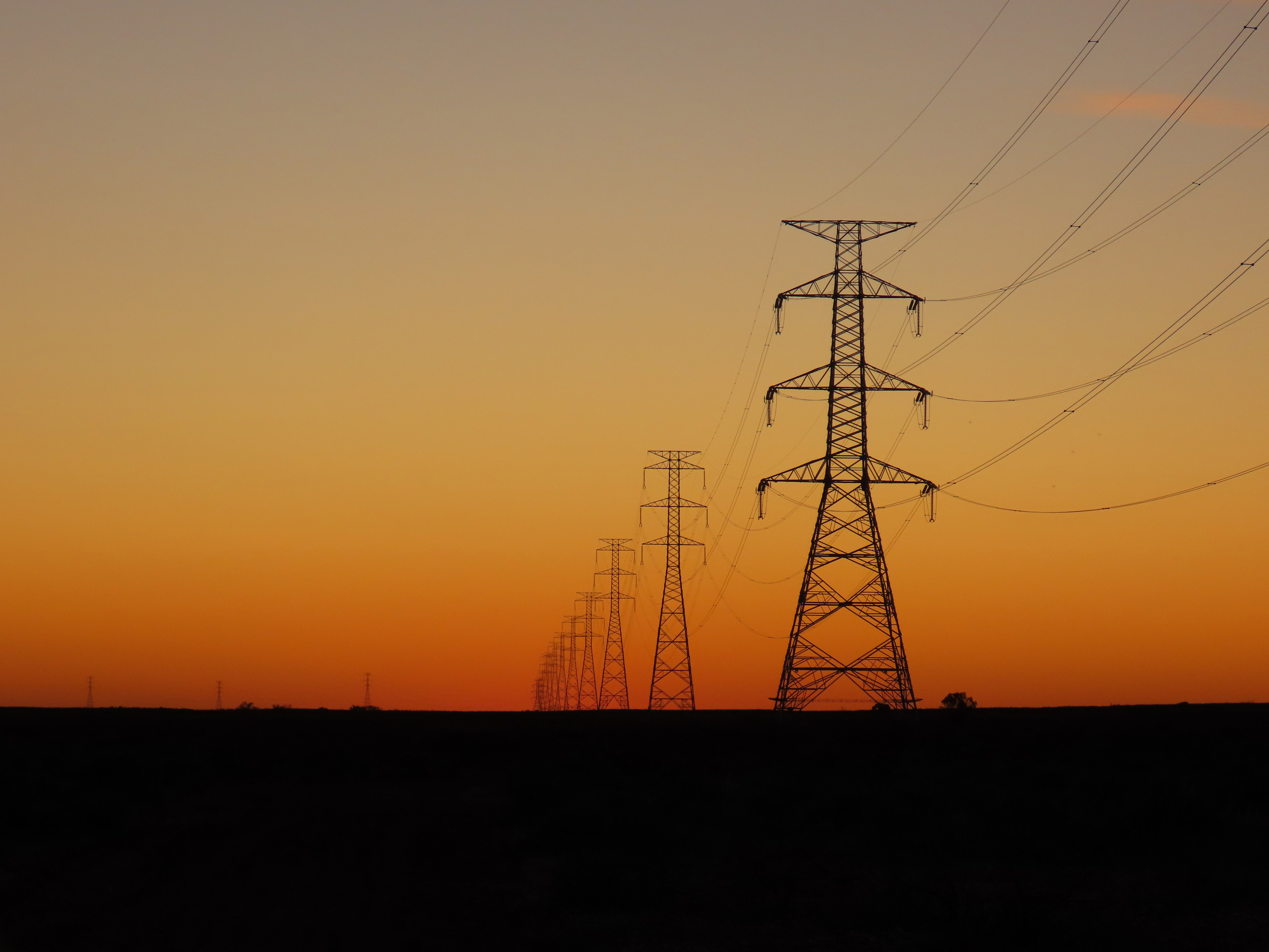 A row of high-voltage power transmission towers stretches into the distance across a flat landscape at sunset. The sky is a gradient of warm orange and soft grey tones, creating a striking silhouette of the towers and power lines against the fading light.