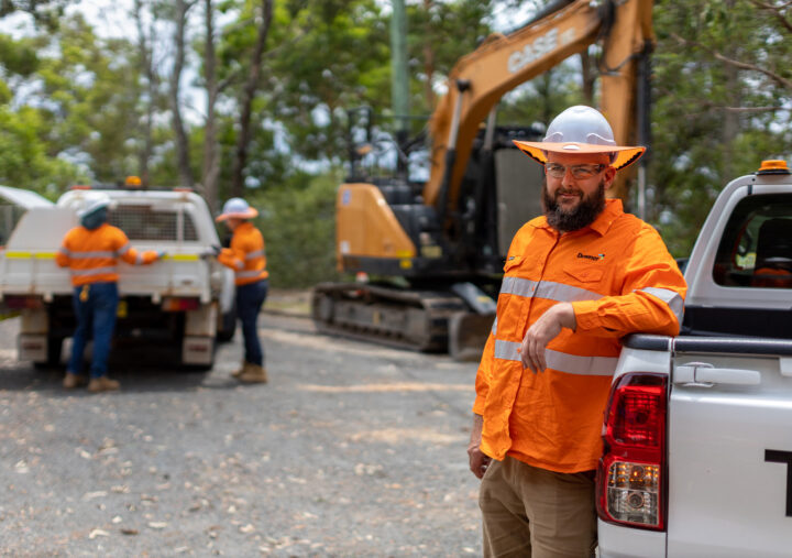 A construction worker wearing a white hard hat, safety glasses, and a high-visibility orange shirt leans against a white utility vehicle on a worksite. In the background, two other workers in similar gear stand near a truck, and an excavator is visible among trees.