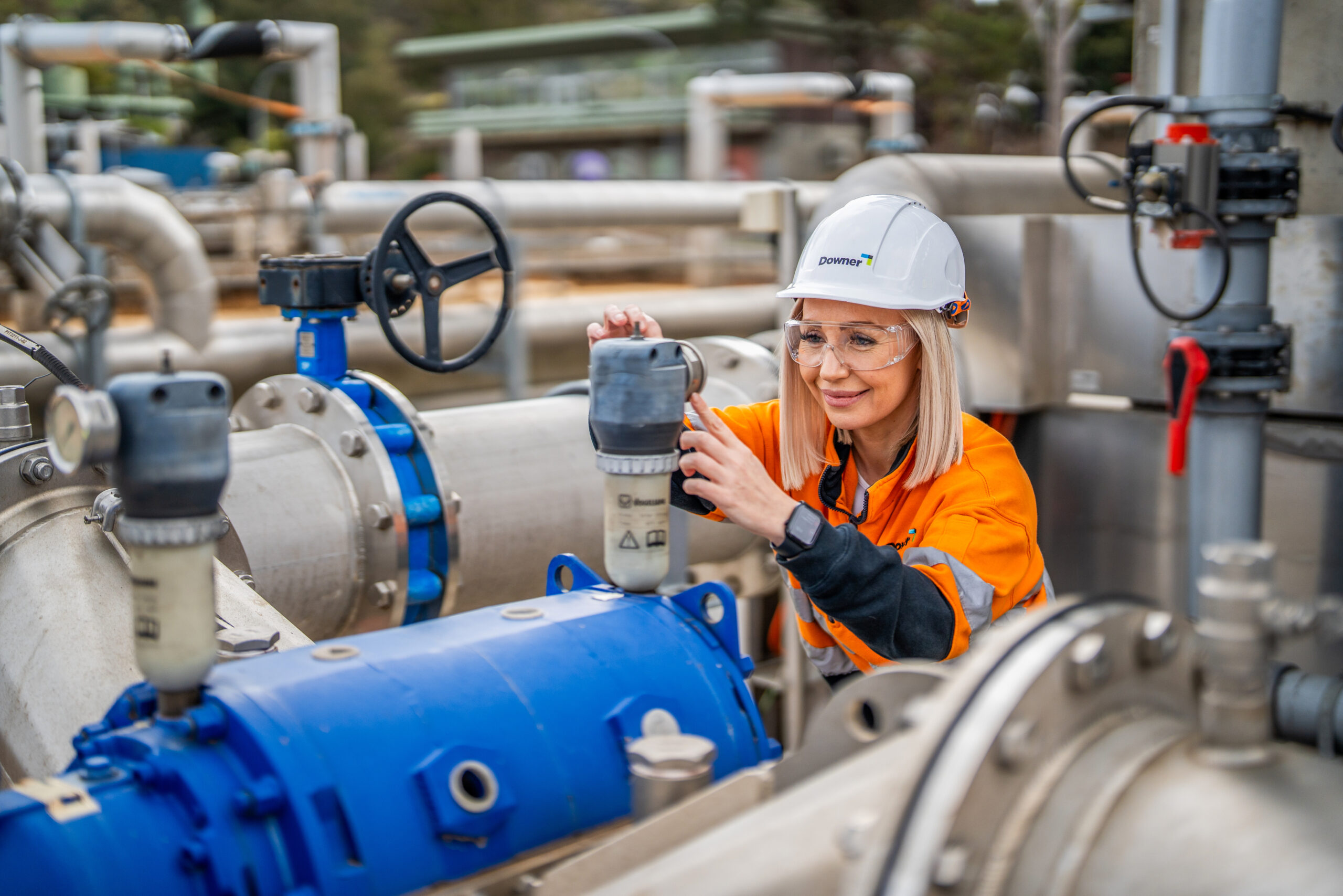 A worker wearing an orange high-visibility jacket, white safety helmet, and protective glasses is adjusting equipment on a large industrial pipe system. The setting is an outdoor facility with metal pipes, valves, and machinery, suggesting maintenance or operations work.