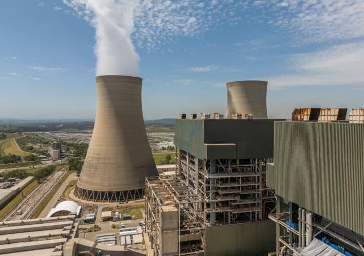 An aerial view of a power generation facility with large cooling towers emitting steam into a partly cloudy sky. Surrounding the plant are industrial buildings, pipes, and infrastructure, set against a landscape of fields and distant hills.