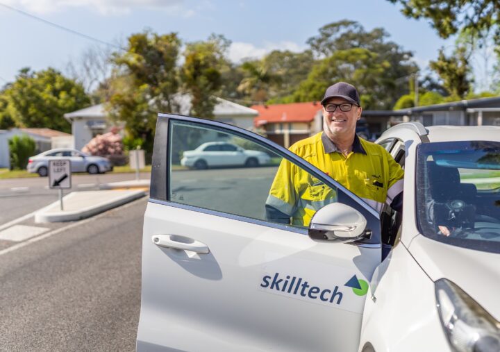 A man in a yellow shirt stands beside a white car, smiling and looking towards the camera.