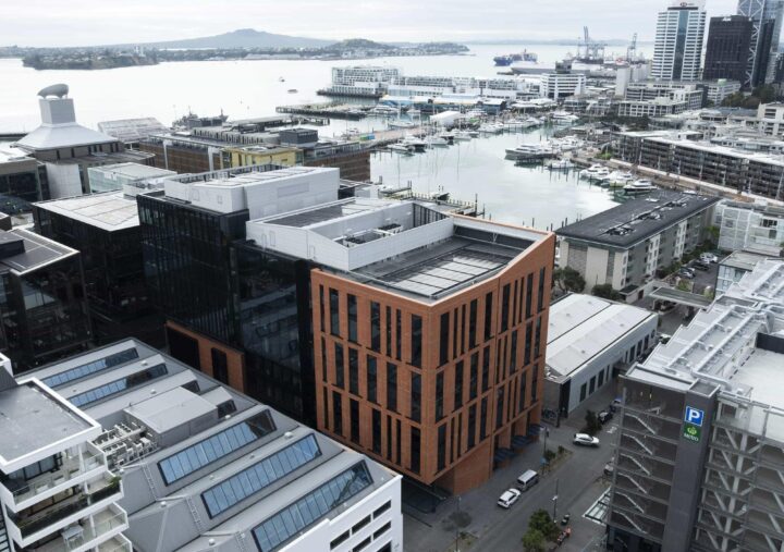 Aerial view of Auckland’s Wynyard Quarter waterfront precinct featuring modern commercial buildings and marinas. In the centre is a prominent mid-rise building with a red brick façade and vertical window patterns, surrounded by glass and metal structures. The harbour and boats are visible in the background under an overcast sky.