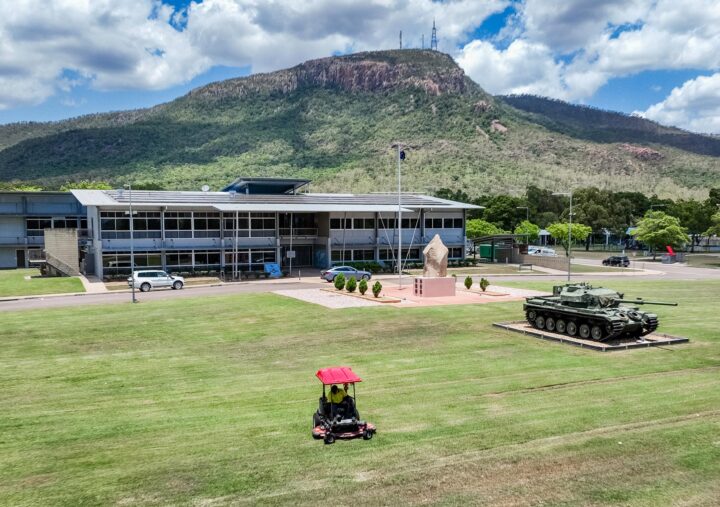 A ride-on mower trims the large lawn outside a Defence facility, where a tank monument and a flagpole stand near the main building. Behind the site, forested mountains rise under a partly cloudy sky.