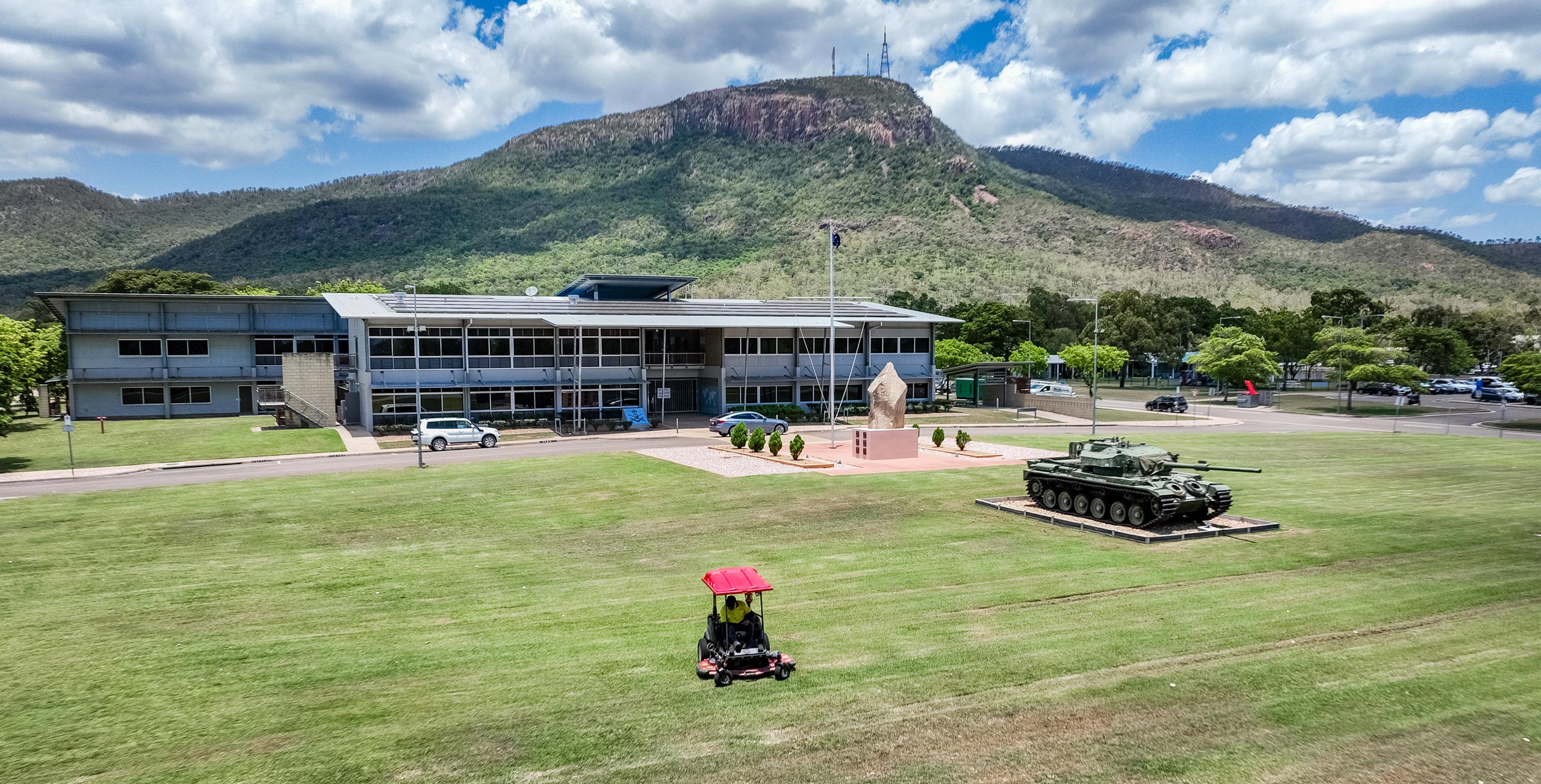 A ride-on mower trims the large lawn outside a Defence facility, where a tank monument and a flagpole stand near the main building. Behind the site, forested mountains rise under a partly cloudy sky.