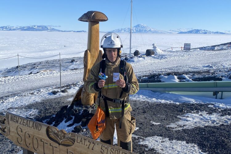 Danny standing at Scott Base sign in his firefighter gear. The New Zealand flag waves proudly in the background.